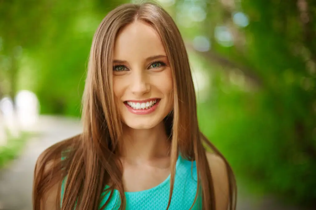 Smiling young woman with long brown hair outdoors in a green park setting, looking hopeful and healthy.
