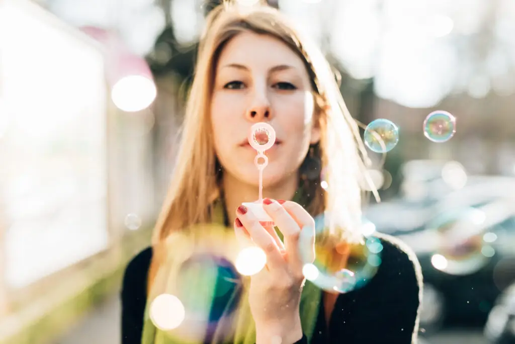 Woman blowing soap bubbles outdoors in soft sunlight, looking peaceful and hopeful.