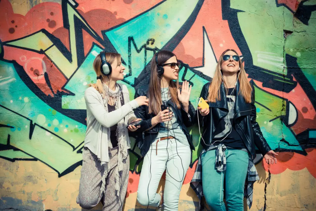 Three young women laughing and listening to music with headphones against a colorful graffiti wall.