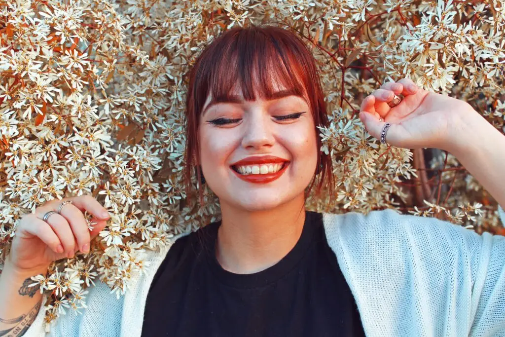 Smiling young woman with red hair lying among white flowers, eyes closed, looking peaceful and hopeful in recovery.