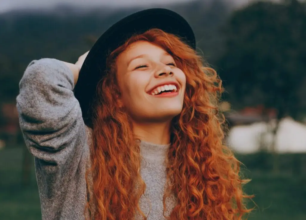 Smiling woman with curly red hair wearing a black hat outdoors, looking joyful and hopeful.