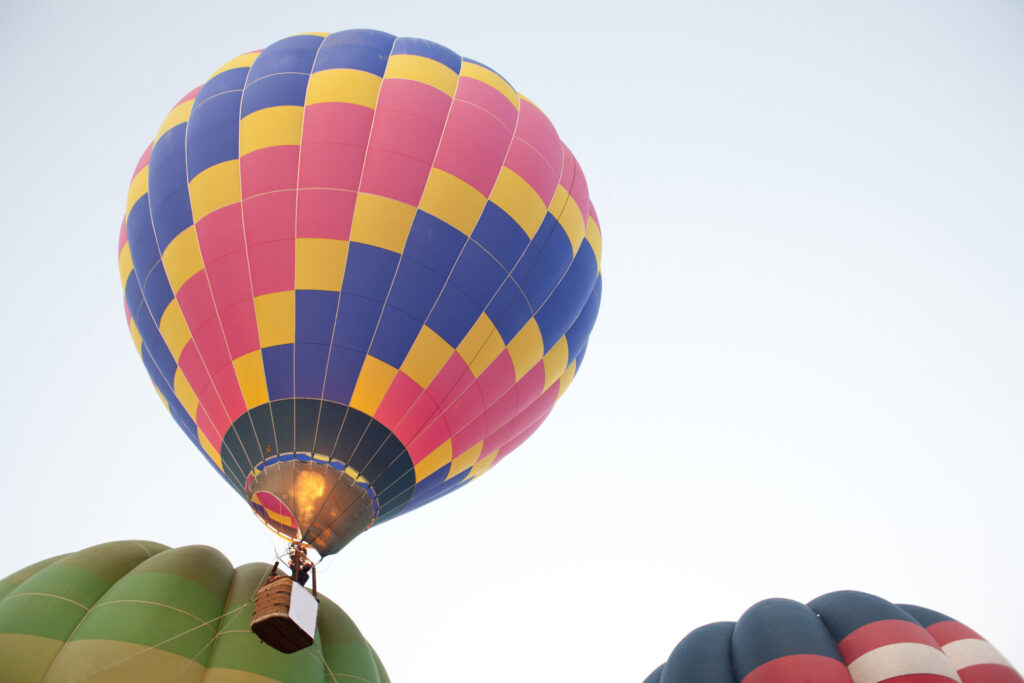 Colorful pink, yellow, and blue hot air balloon rising into a clear sky with burner flame lit, two other balloons below.